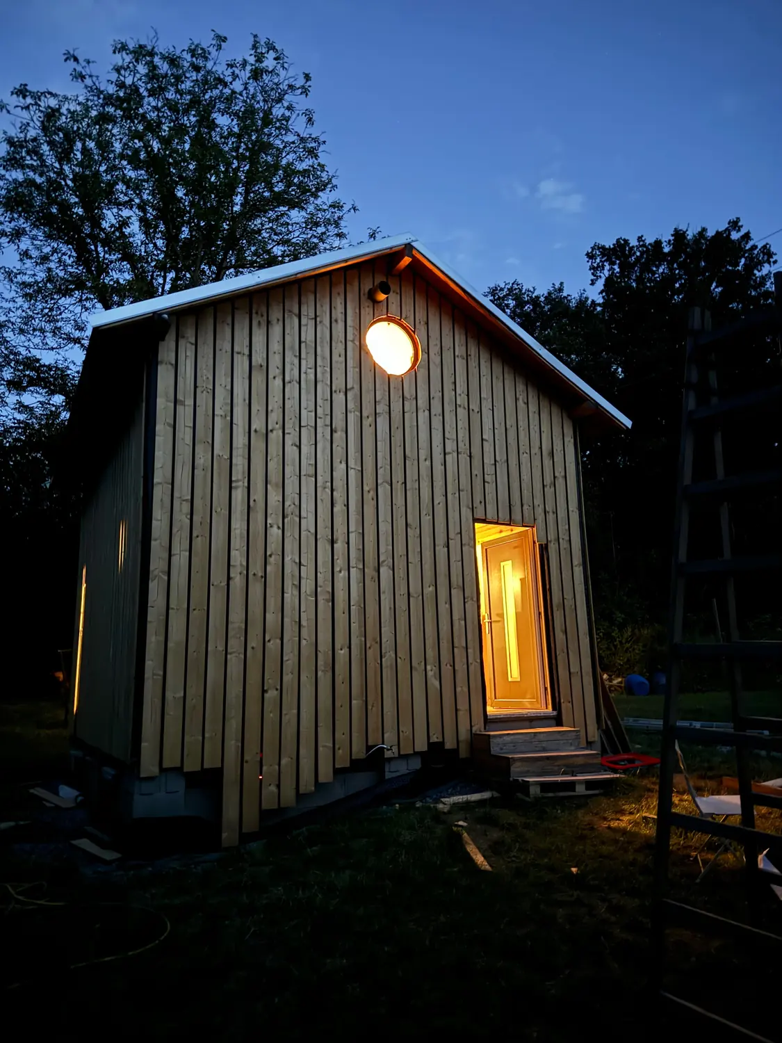 Kleines Holzhaus mit senkrechter Holzverschalung am Abend, Tür beleuchtet